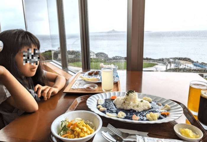沖縄県国頭郡本部町にある美ら海水族館4階のレストラン「イノー」でガラス張りの外（海）を見ながら食事をしている6歳女の子。手前には青い色のカレーとサラダが写っている。